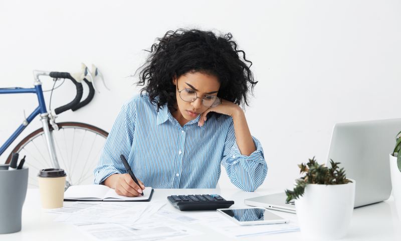 Candid shot of confident hardworking young businesswoman with curly hair sitting at her desk in white office, holding pen while doing accounts by herself, using calculator, laptop and digital tablet