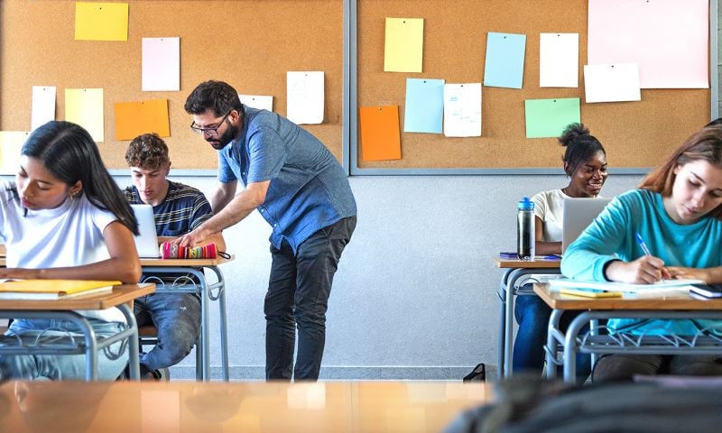 Male teacher helps teen male high school student with lesson in the background. Classmates focused on the task at hand.