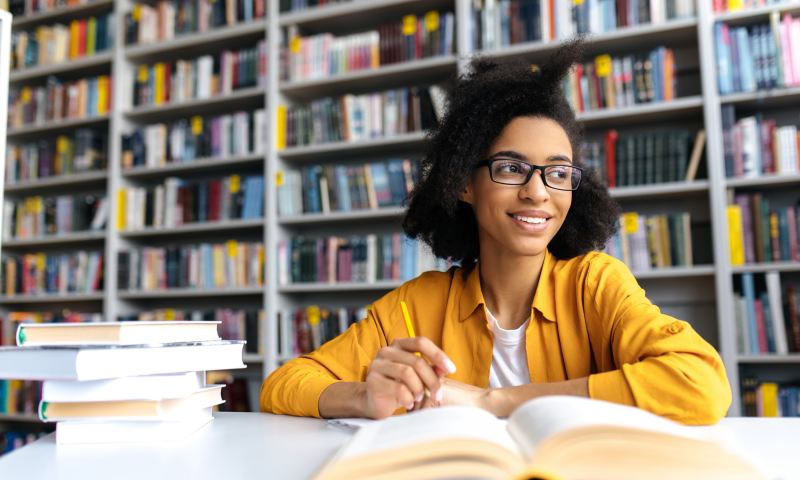 Teenage african american female pretty student with glasses, studying while sits at the table in a college library, reads books to searching information for a lesson or exam, doing homework,looks away